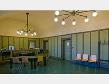 Ein schlicht eingerichteter Raum mit grün gestrichenen Holzwänden, hohen Decken und einem modernen Kronleuchter. Im Vordergrund stehen Tische und Holzstühle, während an einer Wand eine Garderobe und weitere Stühle sichtbar sind; der Raum enthält mehrere Türen.