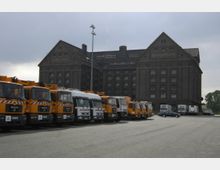 Eine Reihe von orangefarbenen und weißen Lastwagen steht auf einem asphaltierten Platz vor einem großen, dunklen Ziegelgebäude mit Giebeln und der Aufschrift „Zollspeicher“ und „Westhafen“.