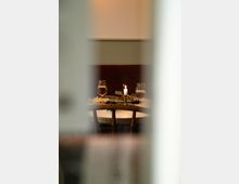 Ein gedeckter Tisch in einem Restaurant mit zwei Weingläsern, einem brennenden Kerzenhalter und Tellern mit Speisen, gesehen durch eine unscharfe Öffnung. Im Hintergrund eine schlichte Wand mit warmem Licht.