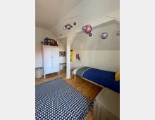 Helles Schlafzimmer im Dachgeschoss mit schrägen Decken, Holzdielen und einem schwarz-weiß gemusterten Teppich. Zu sehen sind ein Einzelbett, ein weißer Kleiderschrank, eine kleine Arbeitsecke sowie ein von der Decke hängendes Planeten-Mobile.