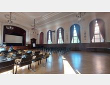 Ein großer Veranstaltungssaal mit hellen Wänden, mehreren hohen, bogenförmigen Fenstern mit Vorhängen und einer Bühne mit rotem Samtvorhang und einer Leinwand. Der Boden ist aus Holz, und der Raum ist bestückt mit mehreren Reihen schwarzer Stühle sowie einem Flipchart an der Seite.