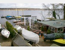 Ein Hafenbereich mit Segelbooten, teilweise mit Planen abgedeckt, an einem Seeufer. Rechts im Bild ein Restaurant mit der Aufschrift "Restaurant Seehase" und einer Terrasse mit leeren Tischen und Stühlen; im Hintergrund Wasser und eine bewaldete Uferlandschaft.