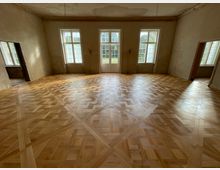 Großer, leerer Innenraum mit hellem, geometrisch gemustertem Parkettboden und hohen Wänden mit teilweise abblätternder Decke. An der Stirnseite befinden sich drei hohe Fenster und eine doppelflügelige Glastür zum Außenbereich, seitlich weitere Tür- und Fensteröffnungen.