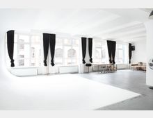 Heller Innenraum eines Lofts mit großen Fenstern, schwarzen Vorhängen, grauem Betonboden und einer weißen, gebogenen Fläche im Vordergrund. Im Hintergrund befinden sich Bürotische, Stühle und eine Sitzgruppe.