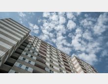 Hochhaus aus Betonplatten mit gleichmäßigen horizontalen Streifen und roten Balkonbrüstungen, aufgenommen aus einer niedrigen Perspektive vor einem Himmel mit vielen kleinen Wolken.