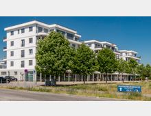 Moderne, weiße Wohn- und Geschäftsanlage mit vier Etagen und großen Fenstern, flankiert von einer Reihe gepflegter, dichter Bäume. Im Vordergrund befindet sich ein blauer Wegweiser zur „Hafenpromenade“ sowie ein Gehweg und eine Straße.