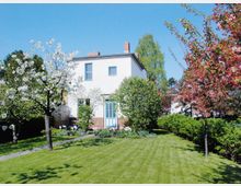 Zweigeschossiges weißes Wohnhaus mit Satteldach, umgeben von einem gepflegten Garten mit grünem Rasen, blühenden Obstbäumen in Weiß und Rosa sowie dichten Sträuchern im Hintergrund. Ein schmaler Weg führt durch den Garten, und der Himmel ist klar und blau.