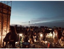 Menschen stehen und sitzen auf einer Dachterrasse bei Dämmerung; im Hintergrund ist die Skyline einer Stadt mit der erleuchteten Silhouette des Berliner Fernsehturms sichtbar. Im Vordergrund befinden sich ein Tisch mit Getränken, Gläsern und Kerzen sowie eine Ziegelsteinwand auf der linken Seite.
