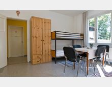 Ein einfacher Raum mit einem Etagenbett, einem Kleiderschrank aus Holz und einem Esstisch mit vier Stühlen. An der Wand ist eine Tür zu einem gelben Flur sichtbar, und große Fenster lassen Tageslicht in den Raum.