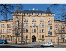 Fassade eines historischen Backsteingebäudes mit symmetrischer Architektur, gekrönten Ecktürmen, spitzbogigen Fenstern und einem zentralen Torbogen. Vor dem Gebäude befinden sich ein Metallzaun, mehrere Flaggenmasten und geparkte Autos, umgeben von kahlen Winterbäumen.