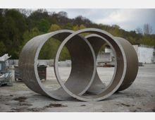 Zwei große Betonringe liegen auf einem offenen, betonierten Lagerplatz. Im Hintergrund sind grüne Bäume, eine weiße Wand und gestapelte Baumaterialien zu sehen.