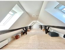 Dachgeschossraum mit hellen, schrägen weißen Wänden und sichtbaren dunklen Holzträgern, ausgestattet mit Fenstern auf beiden Seiten, hellem Holzboden und einzelnen Möbelstücken wie Hockern und Stühlen entlang der Wände.