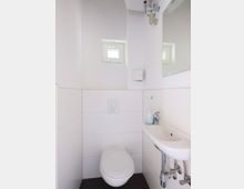 Kleines, modern eingerichtetes Badezimmer mit weiß gefliesten Wänden, einer wandmontierten Toilette, einem kleinen Waschbecken mit Spiegel darüber und einem kleinen Fenster in der oberen Wand.