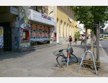 Straßenszene in einer urbanen Umgebung mit einem gelben Gebäude auf der rechten Seite, verzierten Arkadenfenstern und Wänden voller Graffiti. Im Vordergrund steht ein Fahrrad an einem Metallständer, während Fußgänger auf dem Gehweg spazieren und ein Bus im Hintergrund sichtbar ist.
