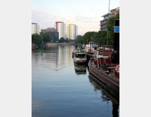 Ein Fluss mit mehreren Anlegestellen, an denen Boote und Schiffe vertäut sind, umgeben von modernen Gebäuden und Wohnhochhäusern in bunten Farben. Im Hintergrund befinden sich grüne Bäume und einige mehrstöckige Gebäude.