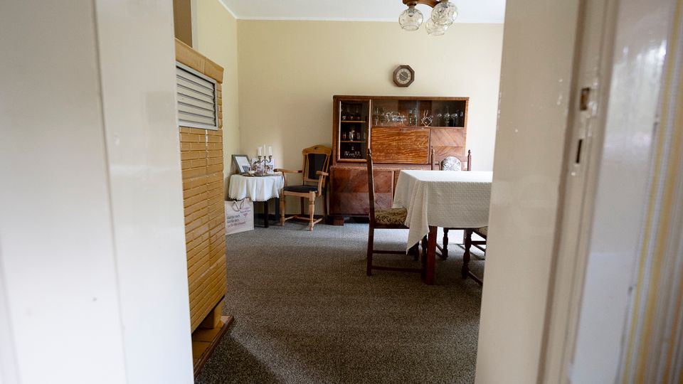 Blick durch eine Tür in ein schlichtes Esszimmer mit grauem Teppichboden, einem Esstisch mit weißer Tischdecke und mehreren Holzstühlen. Im Hintergrund stehen eine dunkle Holzvitrine und ein kleiner Beistelltisch mit Dekoration; an der Decke hängt eine zweiflammige Leuchte.