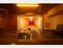 Eine retro-style Kegelbahn mit Holzvertäfelung an den Wänden, einer beleuchteten Decke und zwei Bahnen, die zu beleuchteten Anzeigetafeln führen. Rechts befinden sich eine Holzbank und ein Waschbecken, die Bahnen sind mit einer roten Kugel versehen.