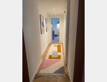 Schmaler, modern eingerichteter Flur mit hellem Holzboden und einem bunten Teppich in geometrischen Mustern. An den weißen Wänden hängen zwei gerahmte Bilder, und der Flur führt zu einem dahinterliegenden, beleuchteten Raum.