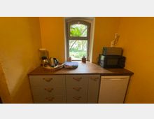 Kleine Küchenzeile mit gelb gestrichenen Wänden, einem Holzfenster mit Blick auf grüne Bäume und einer Arbeitsfläche aus Holz. Darauf befinden sich ein Wasserkocher, ein Mixer, eine Mikrowelle und weitere Küchenutensilien; darunter sind Schubladen und ein Mini-Kühlschrank integriert.