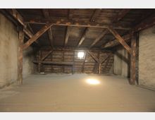 Dachboden mit sichtbarer Holzstruktur, schrägen Wänden und einem kleinen Fenster, das Sonnenlicht in den Raum fallen lässt. Der Raum wirkt leer und zeigt unverputzte Wände sowie einen rohen Holzboden.