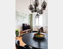 Wohnzimmer mit modernem Design; ein runder, dunkler Holztisch mit einer Schale voller Obst im Vordergrund, eine Hängelampe mit kugelförmigen, spiegelnden Leuchten darüber. Im Hintergrund ein Fernseher auf einem Holzsideboard, ein grüner Vorhang vor einem Fenster und eine dekorative Pflanzenwand.