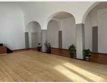 Ein heller Raum mit weiß gestrichenen Wänden und mehreren Rundbögen, die grau umrandete Pfeiler stützen. Der Raum hat einen Holzboden, teilweise hell und teilweise dunkel, und mehrere Zimmerpflanzen stehen verteilt auf dem Boden.