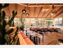 Warm, modern restaurant dining room with long upholstered banquette seating, dark wood tables set with glassware, and pendant lights hanging from a beamed ceiling. Floor-to-ceiling windows overlook trees outside, with potted plants and a large framed wall painting adding decor.