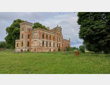 Eine verfallene, mehrstöckige Villa im neoklassizistischen Stil mit roten Ziegel- und Putzfassaden, umgeben von einer großen Wiese mit vereinzelten Bäumen im Hintergrund. Der Himmel ist bewölkt, und ein Metallfass steht auf der Wiese.