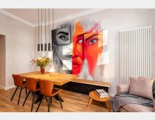 Moderner Ess- und Wohnbereich mit Holztisch, braunen Stühlen und hängenden schwarzen Pendelleuchten, kombiniert mit einem Sofa und kleinem Couchtisch. An der Wand dominiert ein großes, farbiges Wandgemälde mit abstrakten Gesichtern; daneben steht ein hoher, weißer Heizkörper.
