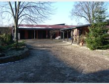 Ein gepflasterter Hof mit Bäumen, Pflanzen und einem Gebäude mit rotem Dach und überdachtem Gehweg. Im Hintergrund sind offene Zugänge und weitere Vegetation sichtbar.