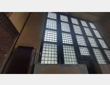 Innenansicht einer Wand mit großen, rechteckigen Fensterflächen aus Glasbausteinen. Links befindet sich ein dunkelroter Heizkörper, und die Wand zeigt teils abblätternde Farbe.