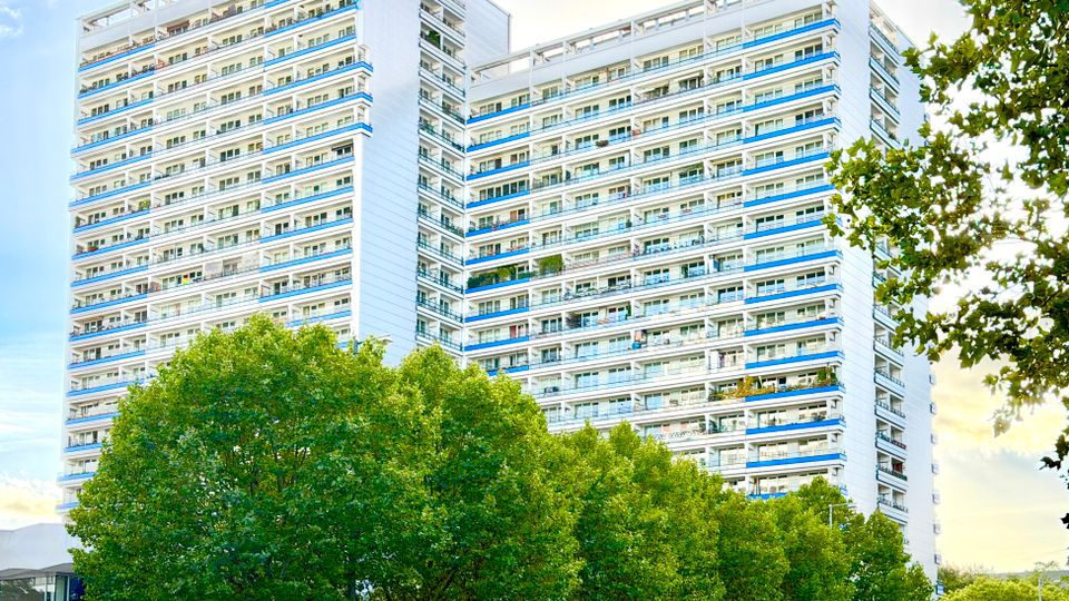 Hochhaus mit weißer Fassade und zahlreichen Balkonen in einem modernen architektonischen Stil, umgeben von grünen Baumkronen im Vordergrund unter einem leicht bewölkten Himmel.