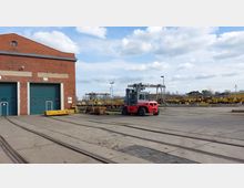 Industriegelände mit einem Backsteingebäude und grünen Rolltoren im Vordergrund. Auf dem Hof stehen ein roter Gabelstapler sowie mehrere gelbe Gleisbau- und Transportfahrzeuge; Schienen verlaufen über den Boden.