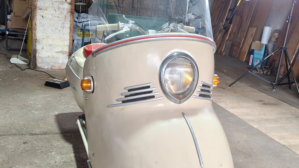 Innenraum einer Werkstatt oder Lagerhalle mit Betonboden, rauen Wänden und gestapelten Holzbrettern sowie Beleuchtungsständer im Hintergrund. Im Vordergrund steht ein kleines, beige lackiertes dreirädriges Kabinenfahrzeug mit großer gewölbter Windschutzscheibe und zentralem Rundscheinwerfer.