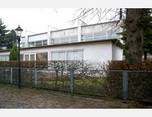 Flaches, modernes Gebäude mit weißen Fassaden, großen Fenstern und einem flachen Dach. Der Vorgarten ist durch eine niedrige Hecke und einen Metallzaun mit Maschendraht abgegrenzt, daneben ein gepflasterter Gehweg und eine Straßenlaterne.