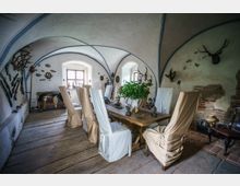Historischer Raum mit gewölbter Decke, einer langen Holztafel und Stühlen mit Hussen. Wände sind mit Jagdtrophäen, Gewehren und rustikalen Dekorationen geschmückt; ein Fenster im Hintergrund lässt Tageslicht herein.
