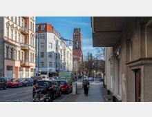 Straßenszene in einer Stadt mit mehrstöckigen Wohngebäuden auf der linken Seite, geparkten Autos entlang der Straße und einer Person auf dem Bürgersteig rechts. Im Hintergrund ist ein roter Kirchturm mit spitzem Dach und einer großen gotischen Fensterfront sichtbar.