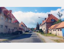 Eine ruhige Straße in einer Wohnsiedlung mit Reihenhäusern im traditionellen Stil, gekennzeichnet durch rote Ziegeldächer und helle Fassaden. Die Umgebung ist von Gehwegen, Bäumen und vereinzelten parkenden Autos geprägt, bei sonnigem Wetter mit blauem Himmel und einigen Wolken.