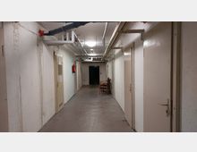 Ein schmaler, karger Flur mit weißen Betonwänden und mehreren geschlossenen Türen in beige auf beiden Seiten. An der Decke befinden sich zahlreiche sichtbare Rohre, und am Ende des Flurs stehen ein paar gestapelte Stühle.