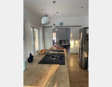 Moderne, helle Wohnküche mit großer Holz-Kücheninsel und integriertem schwarzem Kochfeld, darüber hängende Kugel-Pendelleuchten und eingelassene Deckenstrahler. Links führen große Glas-Schiebetüren mit Vorhängen nach außen, rechts stehen hohe weiße Fronten und ein Edelstahl-Kühlschrank; im Hintergrund öffnet sich der Raum zu einem Wohnbereich.