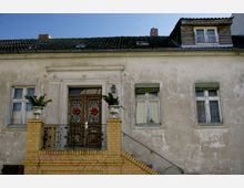 Fassade eines älteren, zweistöckigen Gebäudes mit abgewittertem Putz und dunklem Ziegeldach. Der Eingang wird durch eine Treppe mit gelbem Backstein-Geländer, verzierten Metalltoren und festlicher Dekoration an den Türen hervorgehoben.