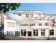 Außenansicht eines modernen Hotels mit der Aufschrift „Inselhotel“ auf der Fassade. Das Gebäude hat eine weiße und rote Backsteinfassade, große Fenster und einen überdachten Eingangsbereich mit einem eleganten, segelförmigen Sonnenschutz.
