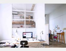 Helles Büro mit großen Fenstern und einer Galerieebene. Im Vordergrund ein Schreibtisch mit Computer, Schreibtischlampen und Arbeitsmaterialien, im Hintergrund Regale, ein Stehpult und eine teilweise geöffnete Tür.