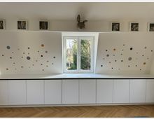 Weiße Dachgaube mit zentralem Fenster, flankiert von Wänden mit bunten Punktmustern in verschiedenen Farben und Größen. Unterhalb des Fensters steht eine weiße Einbauschrankwand mit glatten Fronten auf einem hölzernen Fischgrätboden.