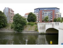 Wohngebäude am Ufer eines Kanals, mit einer modernen roten Backsteinfassade im Vordergrund und älteren Häusern im Hintergrund. Rechts ist eine weiße Brücke mit Verzierungen über das Wasser zu sehen, umgeben von Bäumen und Grünflächen.