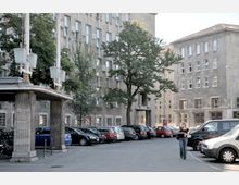 Ein Innenhof mit mehreren mehrstöckigen Gebäuden aus grauem Beton im Hintergrund, umgeben von Bäumen. Im Vordergrund parken zahlreiche Autos, und eine Person läuft über den gepflasterten Platz.