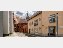 Schmale Gasse mit Kopfsteinpflaster, flankiert von Häusern mit hellen Fassaden rechts und links; im Hintergrund ist eine große, historische Backsteinkirche mit einer grünen Turmspitze zu sehen. Ein Straßenschild mit der Aufschrift „Kirchgasse“ steht rechts im Vordergrund.