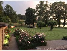 Ein gepflegter Garten mit einem gestreiften Rasen, gesäumt von Rosenbüschen, liegt neben einem kleinen Backsteingebäude. Im Hintergrund sind ein ruhiger Fluss, Bäume und eine beleuchtete Bank zu sehen.
