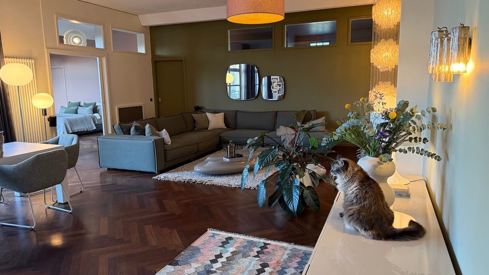 Modern open-plan living room with dark herringbone wood floors, an L-shaped gray sectional around a low oval coffee table, and pendant and wall lights. A white sideboard with vases and a large plant sits beside a geometric rug, with a doorway leading to a bedroom in the background.