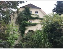 Zweistöckiges Wohnhaus mit hellem Putz und einem dunkelbraunen Satteldach, teils von Efeu bewachsen. Das Gebäude ist von dichtem, grünem Garten mit Bäumen, Sträuchern und hohen Gräsern umgeben.